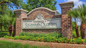The entrance to Savannah Quarters, marked by a beautifully landscaped pathway and a grand clubhouse in the distance. The view of the pristine golf course and tree-lined fairways highlights the luxury and tranquility of this sought-after master-planned community.